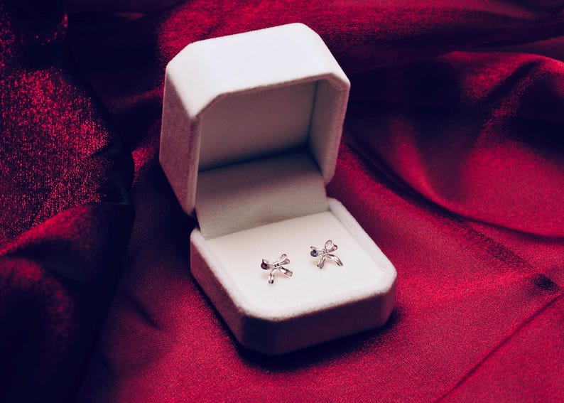 Puede incluir: Un par de pendientes de bot&oacute;n plateados con forma de lazo expuestos en una caja de joyer&iacute;a blanca abierta. La caja descansa sobre una tela de sat&eacute;n rojo intenso, creando una presentaci&oacute;n lujosa. Los pendientes son peque&ntilde;os y delicados.