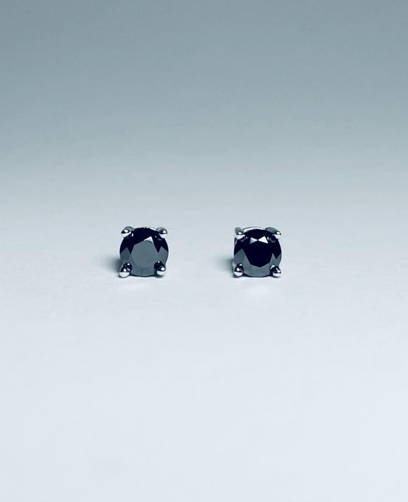 May include: A pair of stud earrings featuring round, black gemstones set in a silver-colored metal. The earrings are displayed against a white background, showcasing their simple elegance and dark sparkle.