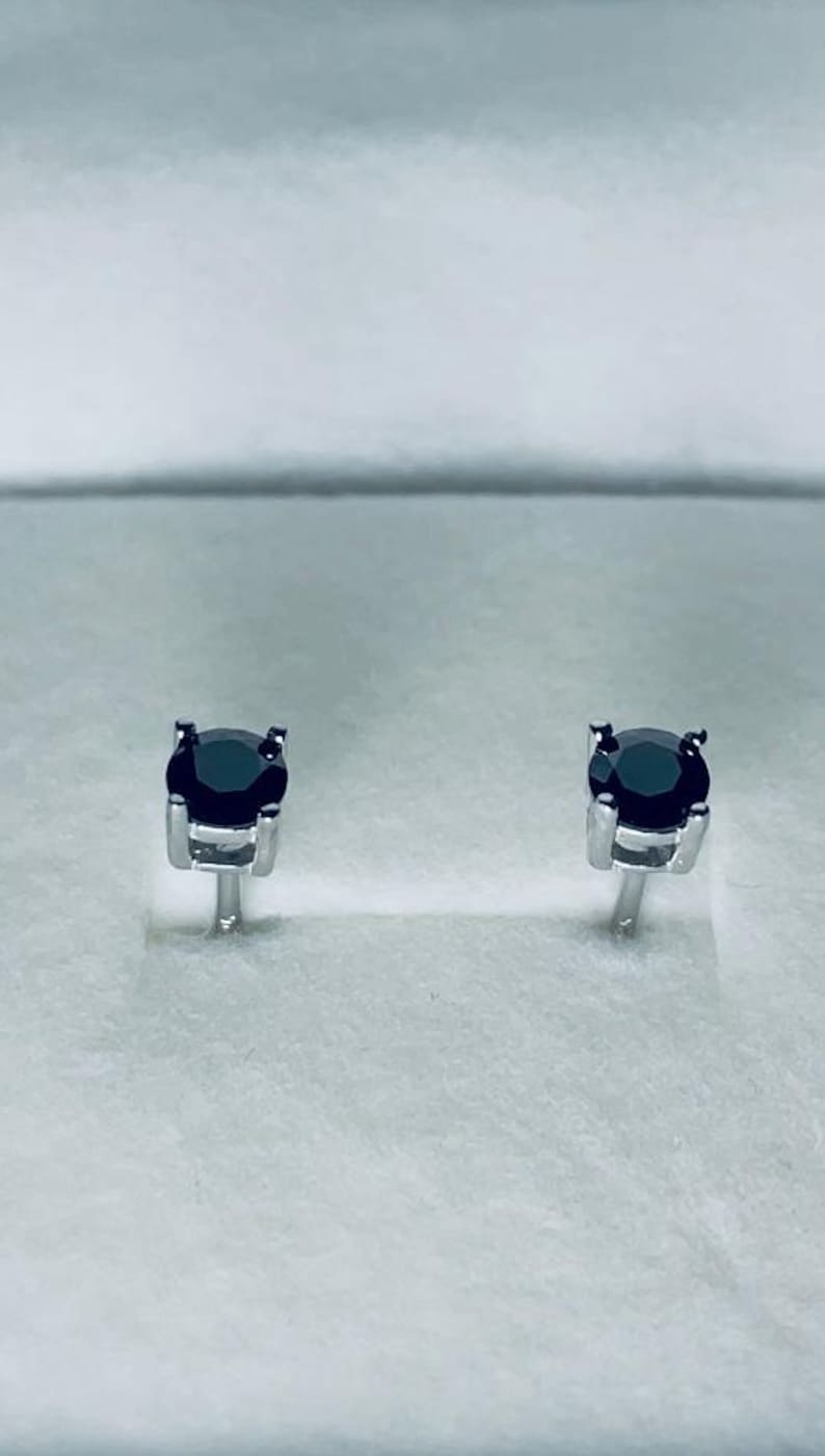 May include: A pair of stud earrings featuring round, black gemstones set in a silver metal. The earrings are displayed on a white surface, showcasing their simple yet elegant design. The gemstones appear to be approximately 0.25 inches in diameter.