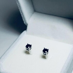 May include: A pair of small, dark purple gemstone stud earrings set in silver metal. The earrings are displayed inside a white, velvet-lined jewelry box with a hinged lid. The box is open, showcasing the earrings.