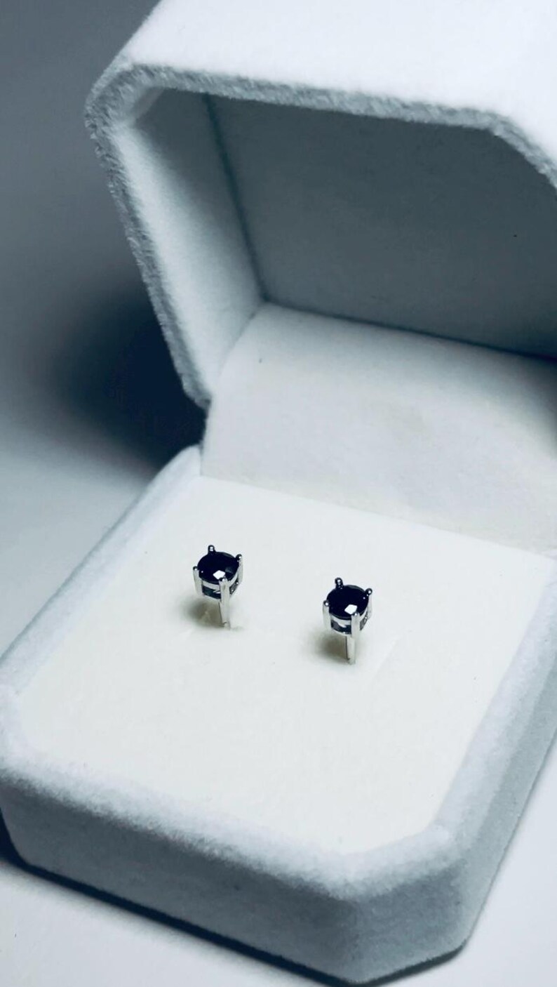 May include: A pair of small, dark purple gemstone stud earrings set in silver metal. The earrings are displayed inside a white, velvet-lined jewelry box with a hinged lid. The box is open, showcasing the earrings.
