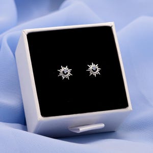 May include: A pair of silver-toned stud earrings shaped like sunbursts with a small dark stone at the center. The earrings are displayed inside a square white gift box with a black velvet insert, featuring a pull-out drawer design.