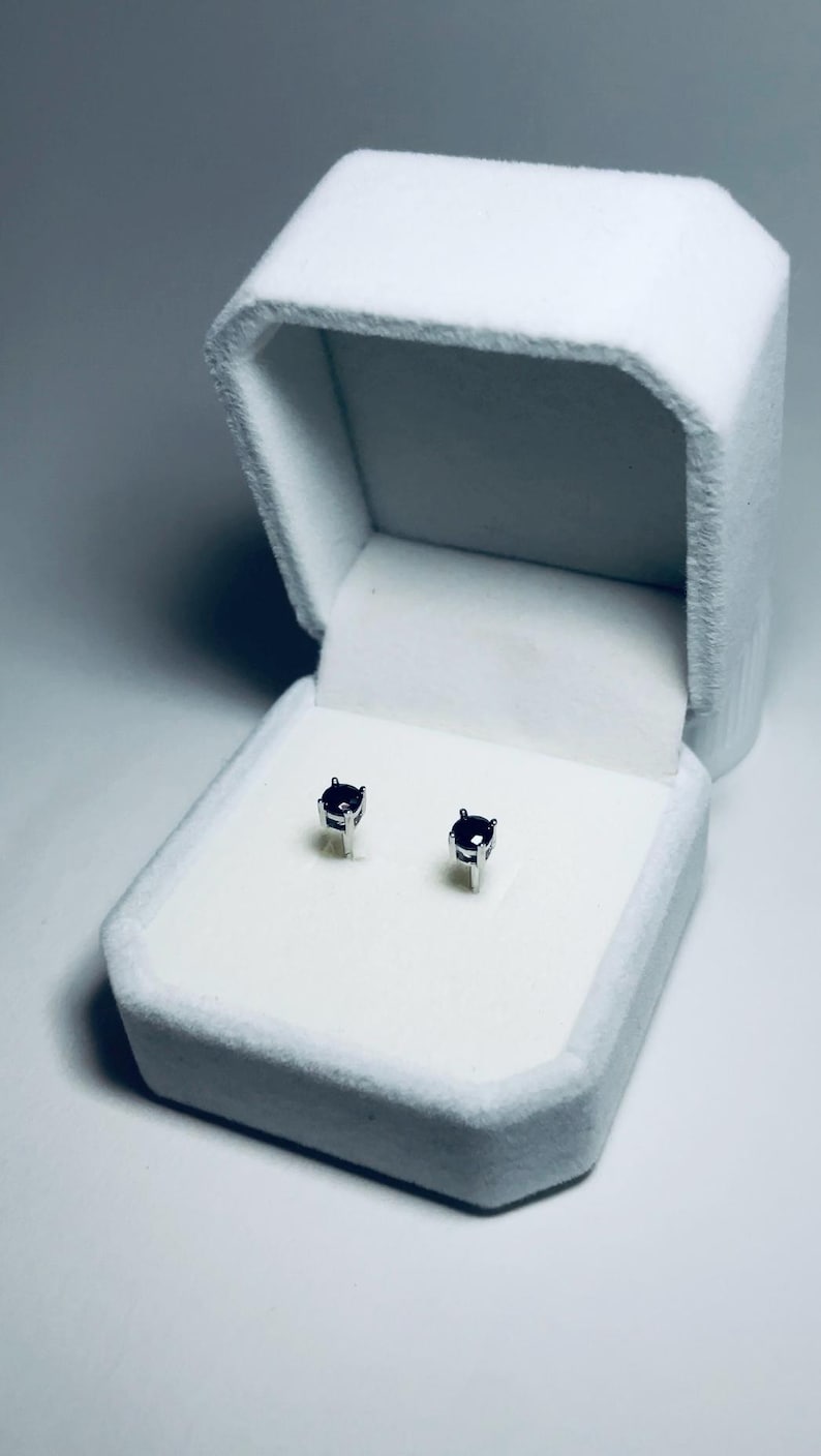 May include: A pair of small, square-cut, dark purple gemstone stud earrings displayed in an open, white velvet jewelry box. The earrings are set in silver-colored metal. The box is rectangular with a hinged lid.