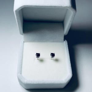 May include: A pair of small, dark blue gemstone stud earrings displayed inside an open, white velvet jewelry box. The earrings are set in silver or white metal. The box is rectangular with a soft, textured interior.