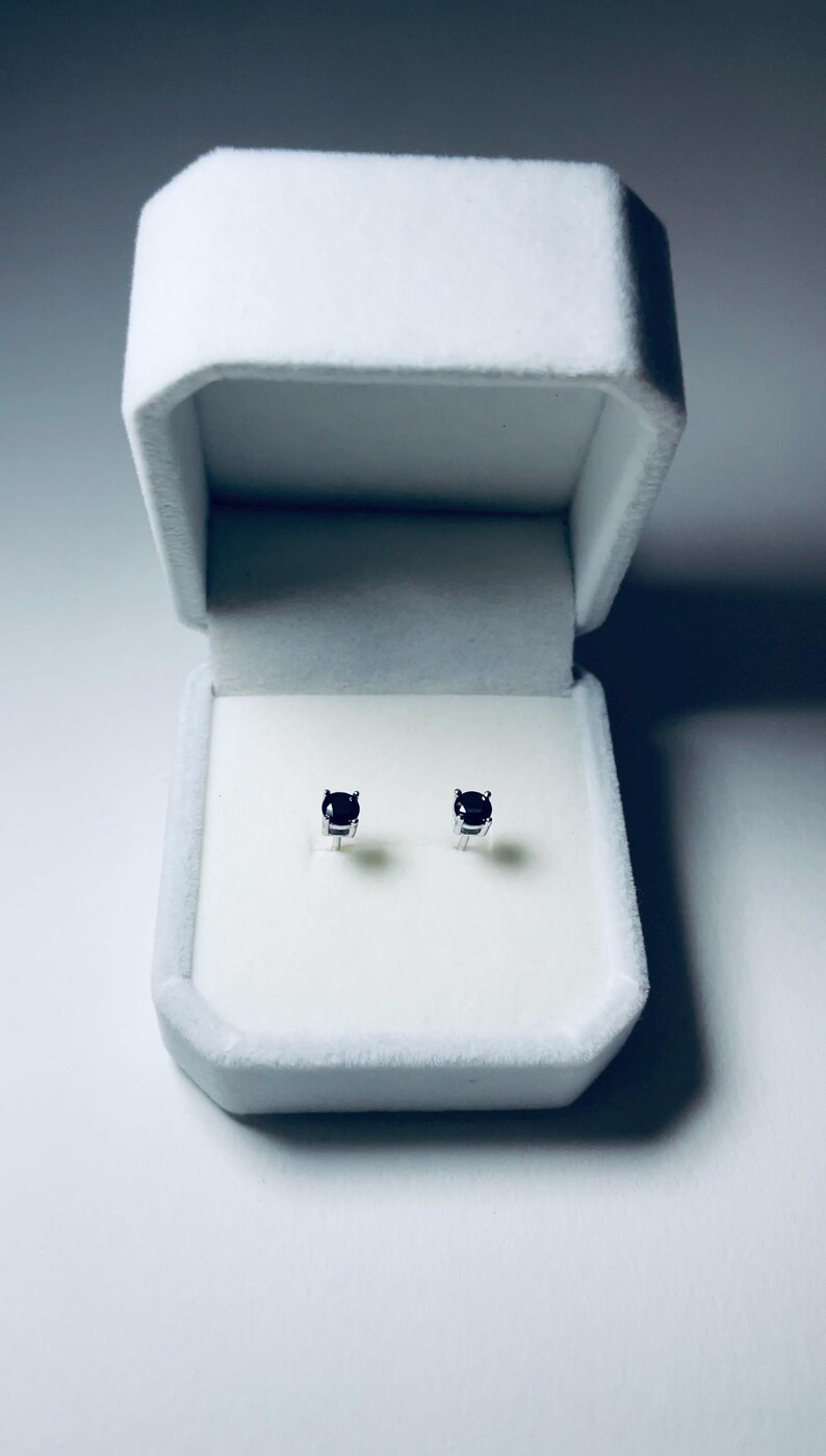 May include: A pair of small, dark blue gemstone stud earrings displayed inside an open, white velvet jewelry box. The earrings are set in silver or white metal. The box is rectangular with a soft, textured interior.