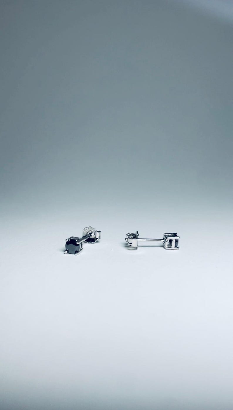 May include: Two pairs of stud earrings. One pair features a black gemstone, the other pair has a clear gemstone. Both pairs have a silver-colored post and backing. The earrings are displayed on a white surface against a light gray background.