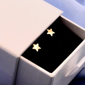 May include: A pair of small, gold-toned star-shaped stud earrings resting inside a white sliding jewelry box. The earrings are positioned on a black velvet insert, contrasting with the white exterior of the box and the light blue fabric.