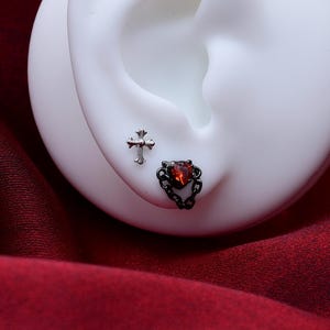 May include: A silver-toned cross stud earring and a black-toned earring featuring a red heart-shaped gemstone surrounded by a delicate metal chain link design, both displayed on a white ear-shaped model for clear visibility of the jewelry.