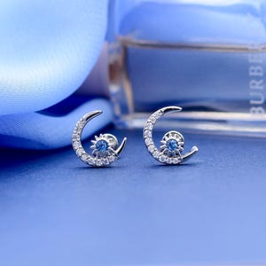 May include: A pair of silver-toned stud earrings shaped like crescent moons, each featuring a small, round, blue gemstone at the center of a starburst design. The crescent moon frames are encrusted with small, clear, sparkling white stones.