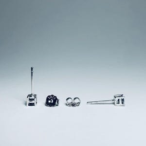 May include: A set of silver stud earrings with a square-cut gemstone. The earrings are displayed with their backs and posts. The earring backs are marked with "S925". The background is a gradient of white to light gray.