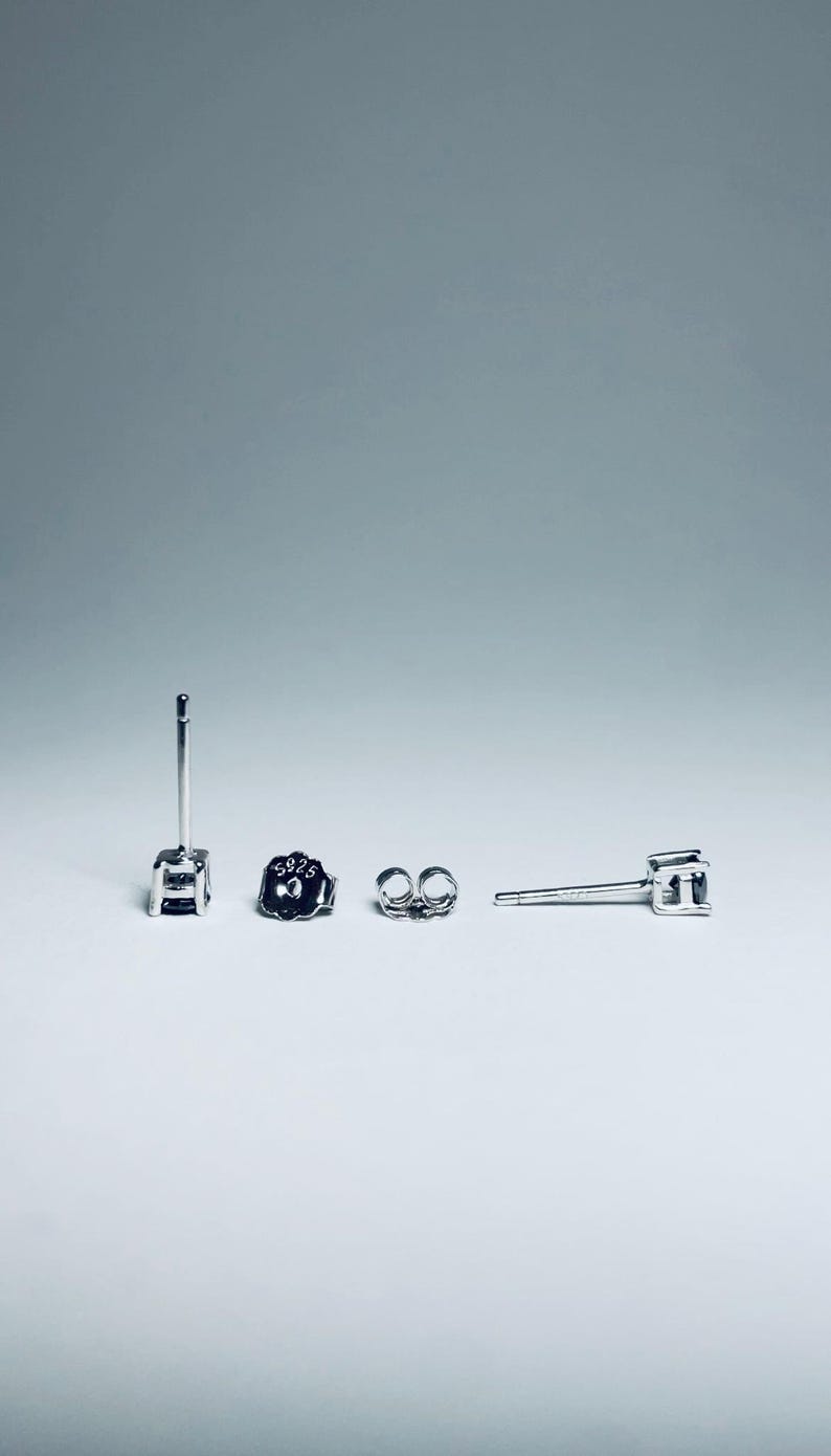 May include: A set of silver stud earrings with a square-cut gemstone. The earrings are displayed with their backs and posts. The earring backs are marked with "S925". The background is a gradient of white to light gray.