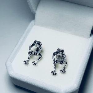 May include: A pair of silver frog-shaped earrings displayed in a white jewelry box. The earrings are detailed with visible legs, eyes, and a textured surface, reflecting light. The frog earrings are a unique accessory.
