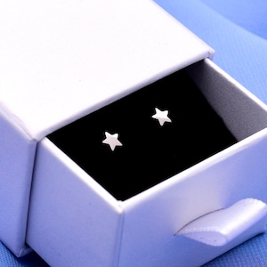 May include: A pair of small, silver-toned star-shaped stud earrings resting inside a white rectangular sliding gift box. The interior of the box is lined with a black velvet insert that holds the two earrings securely in place for display.
