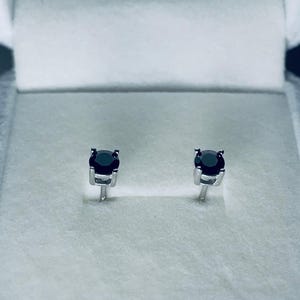 May include: A pair of black gemstone stud earrings, set in silver-colored metal. The earrings are displayed inside a white jewelry box with a soft, textured interior. The gemstones are round and have a dark, reflective surface.