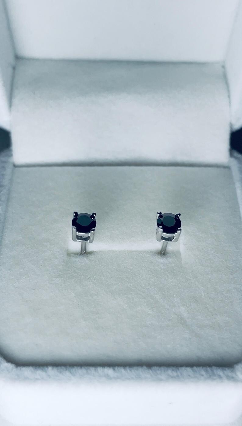 May include: A pair of black gemstone stud earrings, set in silver-colored metal. The earrings are displayed inside a white jewelry box with a soft, textured interior. The gemstones are round and have a dark, reflective surface.