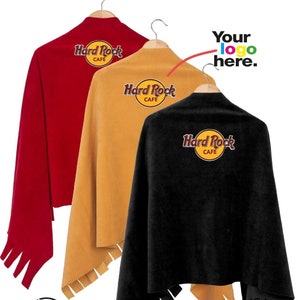 Può includere: Tre coperte in pile con bordi a frangia, appese a grucce. Le coperte sono rosse, dorate e nere. Ogni coperta ha un logo Hard Rock Cafe ricamato sul davanti. Il testo "Your logo here." è scritto sopra le coperte.