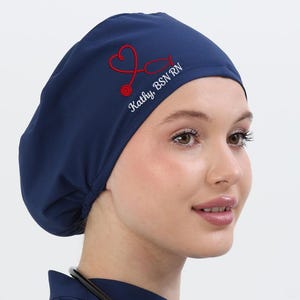 Puede incluir: Un gorro quirúrgico azul marino con un diseño de estetoscopio rojo bordado y el nombre "Kathy BSN RN". El gorro se usa con una parte superior de uniforme azul marino y un estetoscopio negro.