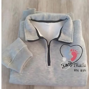 Puede incluir: Sudadera gris con cremallera negra y un corazón con una huella de pie y un estetoscopio. El texto "L&D Thalia RN BSN" está bordado en la sudadera.