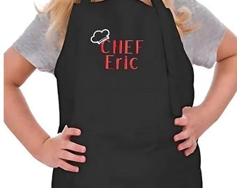 Może przedstawiać: Czarny fartuch dziecięcy z regulowanymi szelkami. Fartuch ma czerwone haftowane napisy "CHEF Eric" z białym rysunkiem czapki szefa kuchni nad tekstem.