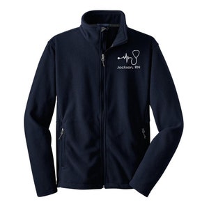 May include: Navy blue fleece jacket with a full zip, two side pockets, and a stand-up collar. A white stethoscope and heartbeat graphic is printed on the chest, with the text "Jackson, RN" below it.