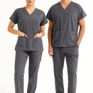 May include: Two people wearing matching gray medical scrubs. The scrubs include a V-neck top with pockets and matching pants. The outfits are made of a soft, wrinkle-resistant fabric. The person on the left is wearing sneakers, and the person on the right is wearing white shoes.
