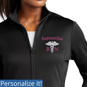 Op de afbeelding: Zwarte jas met ritssluiting en hoge kraag. De jas toont de naam "Samantha" boven een wit en roze medisch symbool met "RN" eronder. De tekst "Personalize It!" staat onderaan de afbeelding.