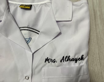 Puede incluir: Bata de laboratorio blanca con cuello y un solo bolsillo. El nombre "Mrs. Alhayek" está bordado en escritura negra en la parte delantera. Un pequeño logotipo es visible cerca del cuello.