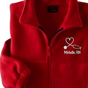Puede incluir: Chaqueta de forro polar roja con cierre de cremallera y un corazón bordado blanco con un estetoscopio y el texto "Michelle, RN".