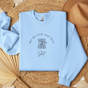 May include: A light blue sweatshirt with a graphic of a coffee maker and the text "Hit me with your best shot".