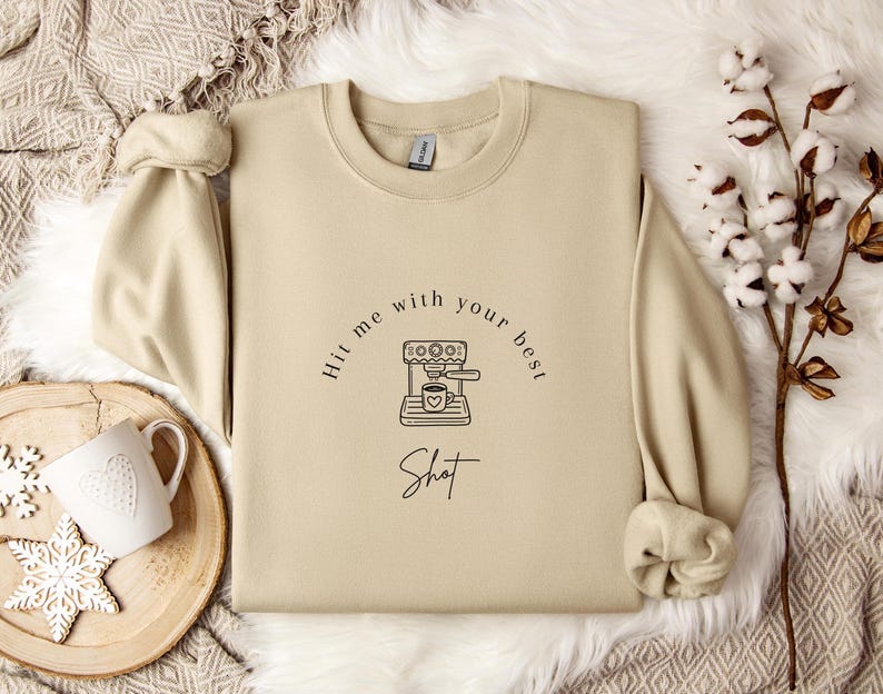 May include: A beige sweatshirt with the text "Hit me with your best shot" and a graphic of a coffee maker with a heart-shaped cup of coffee.