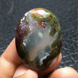 Puede incluir: Un cabujón de ágata verde, marrón y blanco con un centro blanco nublado. La piedra tiene una superficie lisa y pulida.