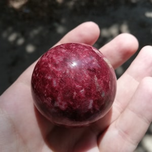 Pode incluir: Uma esfera vermelha lisa e polida com veios brancos e cinzentos. A esfera é feita de pedra natural e tem um acabamento brilhante.