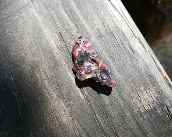 Puede incluir: Un colgante de cristal crudo de color rosa y negro con una anilla de plata.