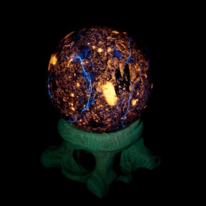 Puede incluir: Un objeto esférico con una superficie oscura y texturizada que presenta patrones de luz naranja y azul, apoyado sobre una base tallada de color verde claro. La esfera muestra una apariencia compleja y moteada con vetas luminosas.