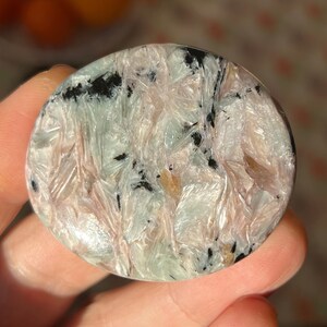 Puede incluir: Una piedra pulida redonda con un patrón único. La piedra presenta una mezcla de colores, incluyendo blanco, verde claro, negro y toques de rosa y beige. La superficie tiene un aspecto texturizado y en capas, con estructuras cristalinas visibles.