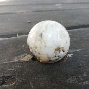 Peut inclure: Une pierre sphérique blanche et grise avec une surface polie. La pierre présente des marques naturelles et de petites imperfections. L'objet est de forme sphérique, possiblement un objet décoratif ou un spécimen naturel.