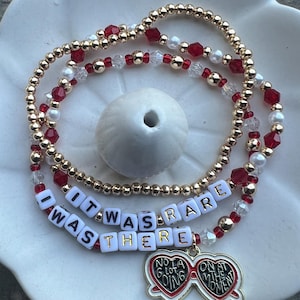 May include: A gold beaded bracelet with red and white beads. The bracelet has a charm that says "It was rare I was there" and a heart-shaped sunglasses charm that says "Not a lot going on at the moment".
