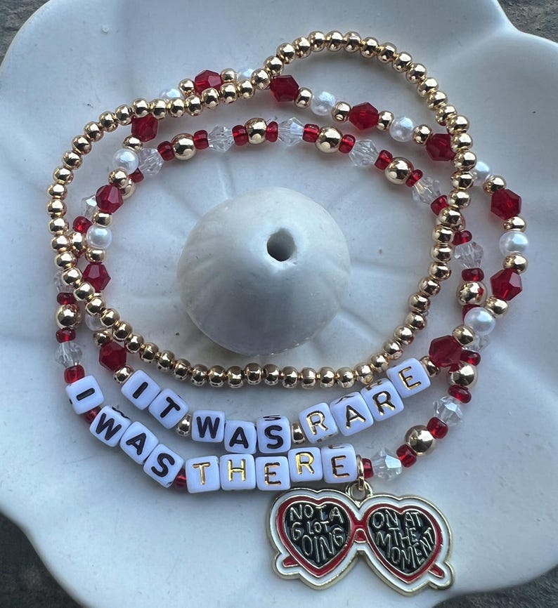 May include: A gold beaded bracelet with red and white beads. The bracelet has a charm that says "It was rare I was there" and a heart-shaped sunglasses charm that says "Not a lot going on at the moment".