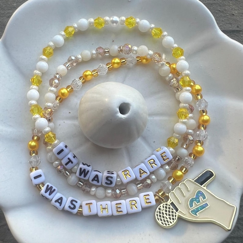 May include: A multi-strand beaded necklace with white and yellow beads. The necklace has a charm with the words "I was there it was rare" and a microphone charm.