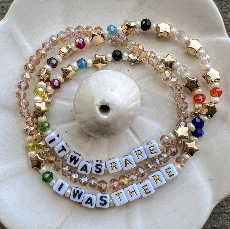 May include: A multi-layered beaded bracelet with gold star charms and white letter beads spelling out the phrase "It was rare I was there".