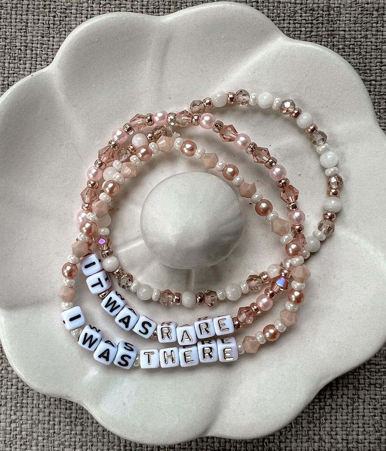 May include: Three layered bracelets with pink and white beads and gold accents. The bracelets spell out the phrase "It was rare I was there".