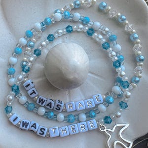 May include: A layered beaded necklace with white and blue beads. The necklace has a silver charm in the shape of a bird and white letter beads that spell out "It was rare I was there".