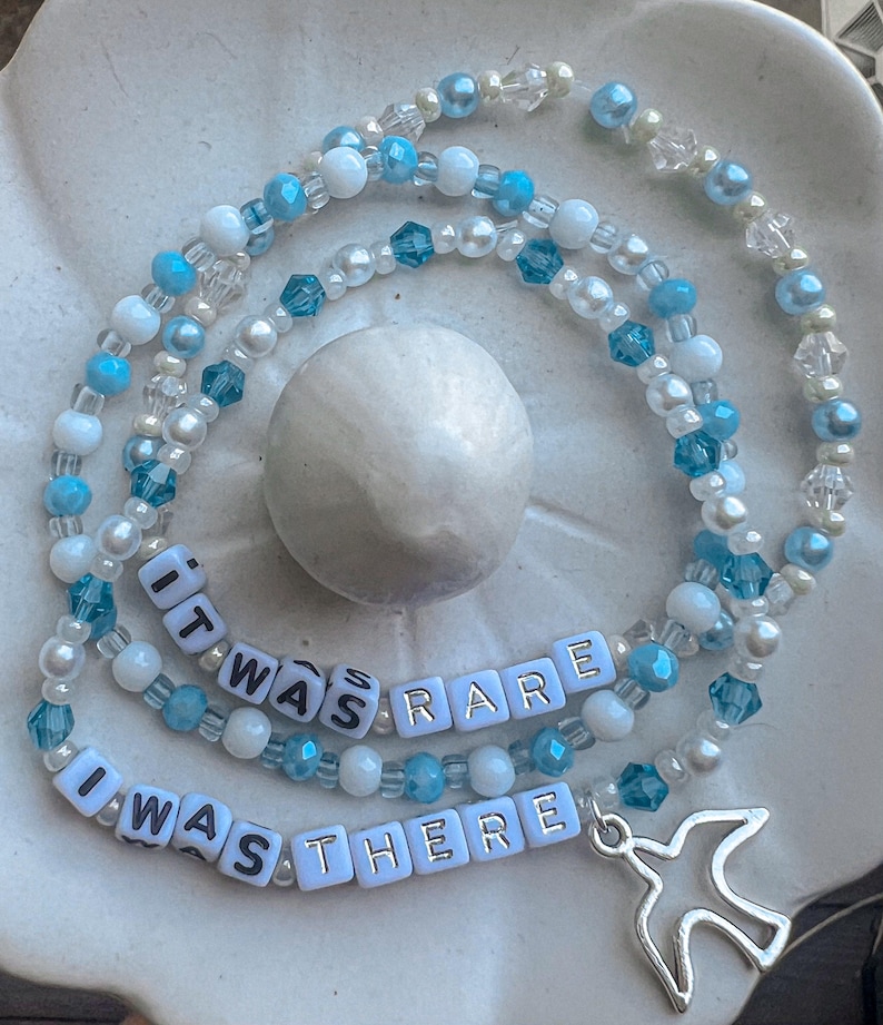 May include: A layered beaded necklace with white and blue beads. The necklace has a silver charm in the shape of a bird and white letter beads that spell out "It was rare I was there".