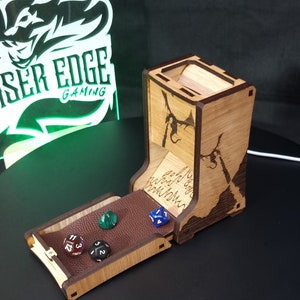 May include: A wooden dice tower with a hinged lid and a brown leather interior. The tower is decorated with a dragon design and the words "And so you shall be known." The tower is open and there are five dice inside, including a blue d6, a green d6, a red d6, a black d6, and a white d6.