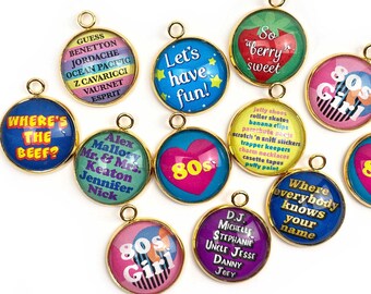 Op de afbeelding: Een verzameling ronde, goudkleurige hangers met diverse kleurrijke ontwerpen en tekst. De hangers tonen zinnen als "Where's the Beef?" en "Let's have fun!" samen met 80s-thema graphics en tekst. De hangers zijn waarschijnlijk voor het maken van sieraden.