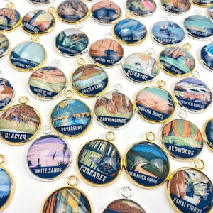 May include: A collection of round, gold-rimmed charms featuring colourful illustrations of various national parks. Each charm has a different park name printed on it, such as "Glacier", "White Sands", "Congaree", and "Redwoods".