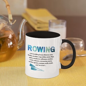 May include: A white ceramic coffee mug with a black rim and handle. The mug has a graphic of a rowing team with the word "ROWING" in blue and white text. The text below the graphic reads "...we're talking boats, blisters, oars, blades, strokes, ergs, slides, seats, flips, splits, workouts, callouses, catches, crabs, releases, recoveries, starts, settles, sprints, sweeps, sculls, wash, and 'let it run'...."