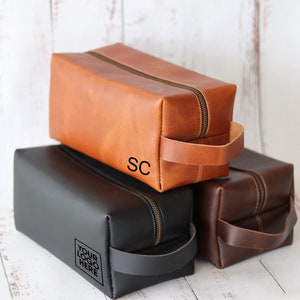 May include: Three leather toiletry bags in brown, black, and dark brown. Each bag has a zipper closure, a handle, and a rectangular shape. The brown bag has the initials "SC" and the black bag has the text "YOUR LOGO HERE".