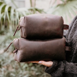 May include: Two brown leather zippered pouches stacked on top of each other. The pouches are made of a soft, textured leather and have a simple, classic design.