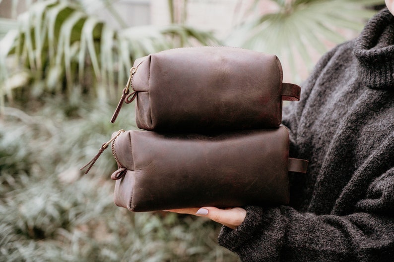 May include: Two brown leather zippered pouches stacked on top of each other. The pouches are made of a soft, textured leather and have a simple, classic design.
